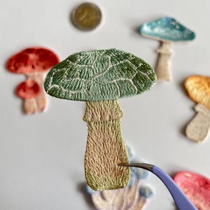 May include: Embroidered mushroom patches in various colors, including green, red, and blue. The patches have detailed stitching and are arranged on a white surface. A pair of tweezers is holding one of the patches.