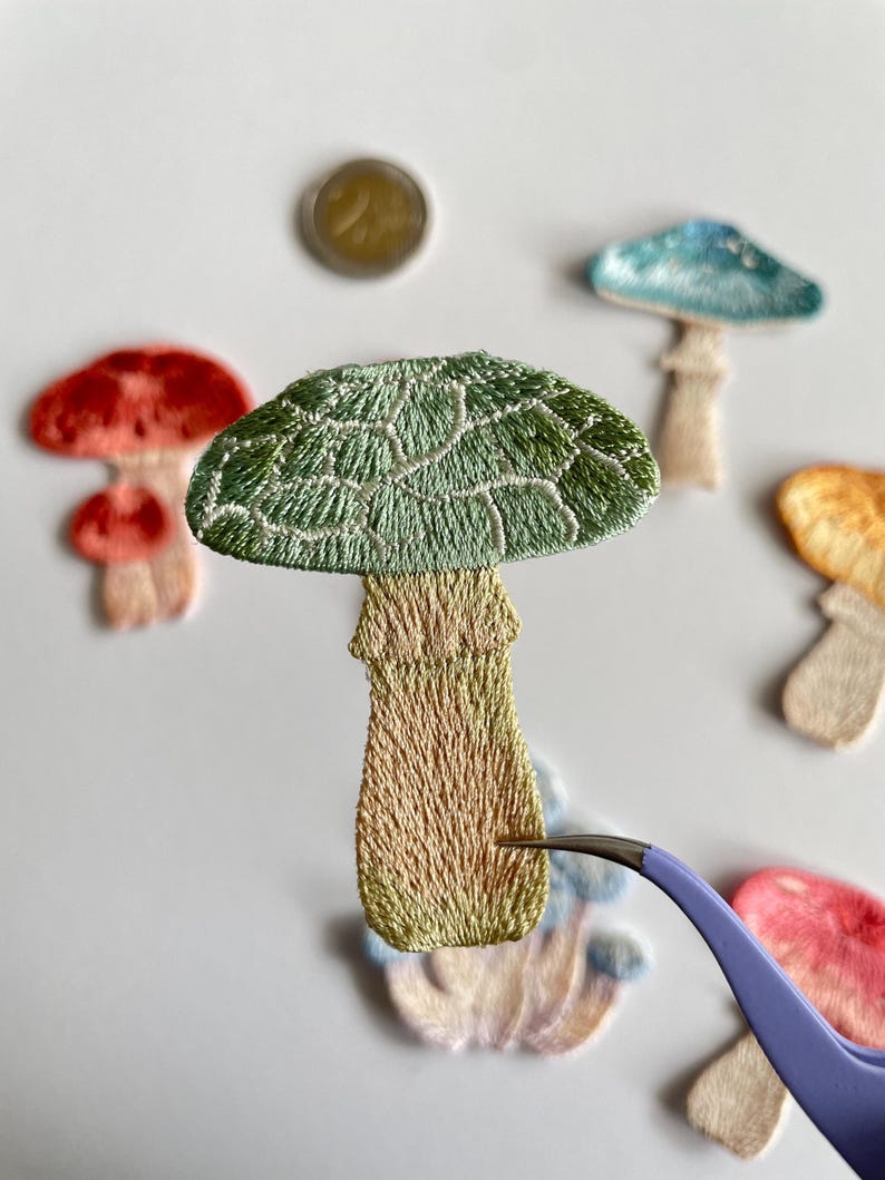 May include: Embroidered mushroom patches in various colors, including green, red, and blue. The patches have detailed stitching and are arranged on a white surface. A pair of tweezers is holding one of the patches.