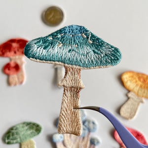 May include: Embroidered mushroom patches in various colors, including teal, red, green, and orange. The patches have detailed stitching and are being held with tweezers. The mushrooms have a whimsical, handmade aesthetic.