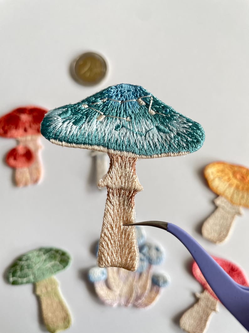 May include: Embroidered mushroom patches in various colors, including teal, red, green, and orange. The patches have detailed stitching and are being held with tweezers. The mushrooms have a whimsical, handmade aesthetic.