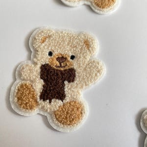 Pode incluir: Patches bordados de ursinho de pel&uacute;cia em creme, castanho e bege. Os patches apresentam um design de ursinho de pel&uacute;cia com um cachecol castanho. Os patches est&atilde;o em um fundo branco. Ideais para roupas ou acess&oacute;rios.