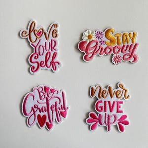 Può includere: Quattro toppe ricamate con messaggi positivi. Le toppe presentano le frasi "Love Your Self", "Stay Groovy", "Be Grateful" e "Never Give Up" in rosa, oro e bianco.
