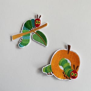 Könnte beinhalten: Zwei gestickte Aufnäher mit einer grünen und blauen Raupe. Eine Raupe sitzt auf einem Ast mit einem Blatt, die andere auf einem orangefarbenen Apfel. Die Raupe hat einen roten Kopf und lächelt.