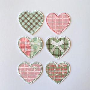 May include: Six heart-shaped embroidered patches arranged in two columns. The designs include a green and brown gingham pattern, a pink grid, a split red and green plaid, a green plaid with a white bow, a pink grid with a small bow, and a green plaid with white stars.