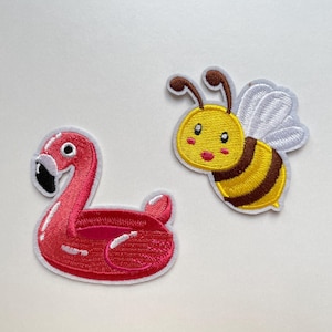 Könnte beinhalten: Zwei gestickte Aufnäher: ein pinkfarbener Flamingo-Pool-Schwimmer und eine gelb-braune Cartoon-Biene mit weißen Flügeln. Der Flamingo hat einen schwarzen Schnabel und einen weißen Rand. Die Biene hat ein lächelndes Gesicht und braune Fühler.