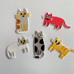 Könnte beinhalten: Fünf gestickte Katzenaufnäher in verschiedenen Farben und Mustern. Das Set enthält eine hellgraue Katze mit orangefarbenen Flecken, eine einfarbige leuchtend rote Katze, eine weiße Katze, eine graue Katze mit schwarzen Flecken und eine gelbe Katze mit orangefarbenen Streifen.