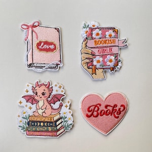 Könnte beinhalten: Vier bestickte Aufnäher mit einem rosa Buch mit Schleife und dem Wort Love, einem mit weißen Blumen verzierten Buch mit der Aufschrift Bookish Girlie, einem rosa Drachen auf einem Bücherstapel und einem herzförmigen Aufnäher mit dem Wort Books.