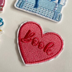May include: A pink heart-shaped embroidered patch with the word "Love" in red cursive. The patch has a white border and is on a white background. Other patches are also visible in the image.