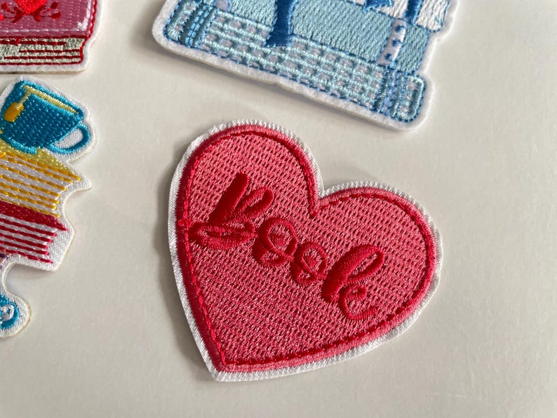 May include: A pink heart-shaped embroidered patch with the word "Love" in red cursive. The patch has a white border and is on a white background. Other patches are also visible in the image.