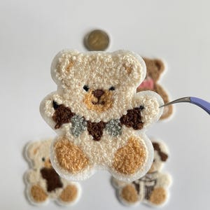 Pode incluir: Close-up de v&aacute;rios patches de ursinhos de pel&uacute;cia. O patch principal &eacute; um urso creme com detalhes marrons no nariz, patas e uma gola decorativa. Outros patches incluem um urso marrom e um urso com um cachecol. Os patches est&atilde;o em um fundo branco.