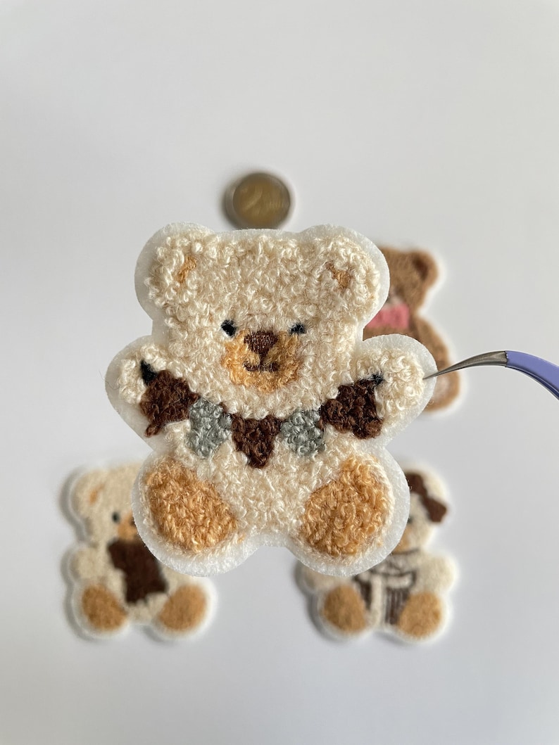 Pode incluir: Close-up de v&aacute;rios patches de ursinhos de pel&uacute;cia. O patch principal &eacute; um urso creme com detalhes marrons no nariz, patas e uma gola decorativa. Outros patches incluem um urso marrom e um urso com um cachecol. Os patches est&atilde;o em um fundo branco.