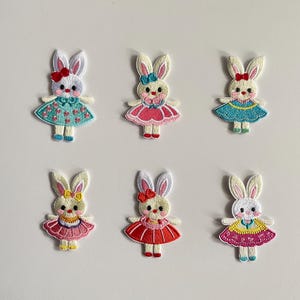 Puede incluir: Seis parches bordados de conejitos en varios colores. Cada conejito tiene pelaje blanco, orejas largas y un lazo. Llevan vestidos en tonos rosa, azul y amarillo. Los parches son adecuados para coser en ropa o accesorios.