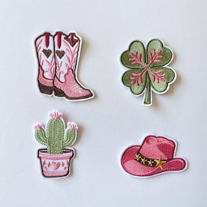 Op de afbeelding: Een set van vier geborduurde patches met een paar wit-roze cowboylaarzen met hartdetails, een groen klavertjevier met roze nerven, een groene cactus in een roze pot en een roze cowboyhoed met een gouden sterrenband.