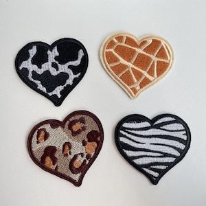 May include: Four heart-shaped embroidered patches with animal print designs. The patterns include cow, giraffe, leopard, and zebra prints. Each patch has a black border and is set against a white background.