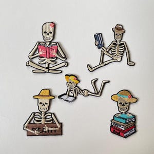 Könnte beinhalten: Fünf gestickte Aufnäher mit Skeletten in verschiedenen Posen mit Büchern. Ein Skelett sitzt im Schneidersitz mit einem rosa Buch, ein anderes mit einem blauen Buch und Hut, eines liegt mit einem gelben Hut, eines hält ein braunes Schild und eines sitzt auf Büchern.
