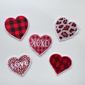 Può includere: Cinque toppe a forma di cuore con bordi bianchi. I disegni includono quadri rossi e neri, stampa leopardata rosa, un cuore rosa con ricamo "XOXO", un cuore rosa con ricamo "LOVE" e un motivo a rombi rosso e nero.