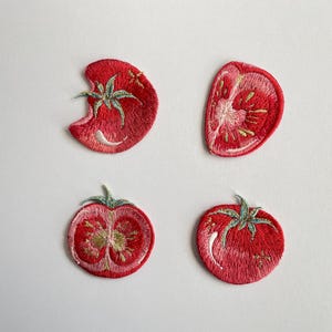 Könnte beinhalten: Vier bestickte Aufnäher mit Tomatenmotiven in Rot-, Rosa- und Grüntönen. Das Set besteht aus zwei ganzen Tomaten mit Stielen und zwei aufgeschnittenen Tomatenhälften, die alle mit detaillierter Stickarbeit und sichtbaren Texturen versehen sind.