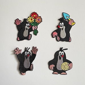 Könnte beinhalten: Vier aufgestickte Aufnäher mit einer Cartoon-Maulwurf-Figur. Der Maulwurf ist schwarz mit rosa Händen und Füßen und einem weißen Bauch. Ein Aufnäher hält Blumen, ein anderer eine Schaufel, ein anderer hat die Arme erhoben, und der letzte hält einen Ball.