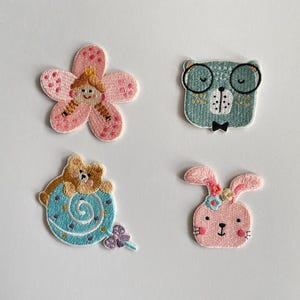 Könnte beinhalten: Vier gestickte Aufnäher mit Cartoon-Designs. Eine rosa Blume mit einer Prinzessin, ein tealfarbener Bär mit Brille und Fliege, ein Bär auf einer blauen Spirale und ein rosa Hase mit Blumen.