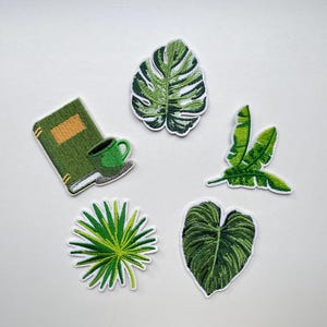 Op de afbeelding: Geborduurde patches met een groen boek, een koffiekopje en diverse tropische bladeren. De patches zijn in groen- en wittinten, met gedetailleerde stiksels. Perfect om kleding of accessoires mee te personaliseren.