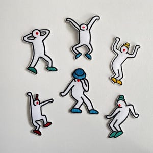 Könnte beinhalten: Sechs gestickte Aufnäher mit weißen Strichmännchen in verschiedenen Tanzposen. Jede Figur hat einen roten Punkt als Gesicht und trägt farbige Accessoires wie Mützen und Schuhe in Blau-, Grün-, Gelb- und Rottönen mit schwarzen Umrandungen.