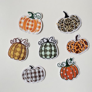 Puede incluir: Colección de siete parches de calabaza bordados en varios patrones y colores. Los diseños incluyen cuadros naranjas, estampado de leopardo y patrones de diamantes verdes. Cada parche tiene un borde blanco y un tallo verde.