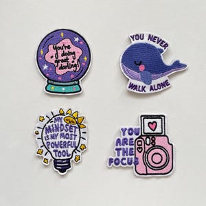 May include: Four embroidered patches with inspirational quotes. One patch is a crystal ball with the text "You're doing great darling!". Another is a whale with the text "You never walk alone". A third is a lightbulb with the text "My mindset is my most powerful tool". The last is a camera with the text "You are the focus".
