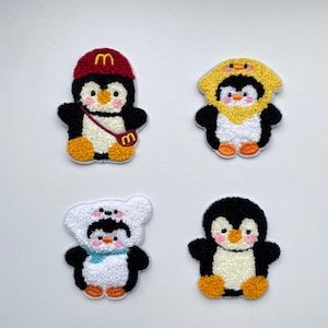 Könnte beinhalten: Vier gestickte Pinguin-Patches in verschiedenen Outfits. Einer trägt eine rote Mütze und eine Tasche mit goldenem "M"-Logo. Ein anderer trägt ein gelbes Kapuzen-Outfit. Der dritte ist in einem weißen Bärenkostüm, und der letzte ist ein klassischer Pinguin.