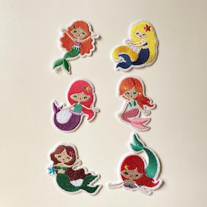 May include: A set of six embroidered mermaid patches featuring various hair colors including orange, yellow, pink, red, and brown. Each mermaid has a distinct tail color, such as green, blue, purple, or pink, and small starfish hair accessories.
