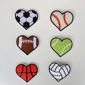 May include: Six heart-shaped embroidered patches featuring sports balls: soccer, baseball, football, tennis, basketball, and volleyball. Each patch has a black border and is designed with the respective sport's details, perfect for crafting or personalization.