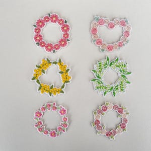 Puede incluir: Seis parches florales bordados en varias formas de corona. Los parches presentan flores rosas, amarillas y verdes. Un parche tiene forma de corazón. Los parches están sobre un fondo blanco.