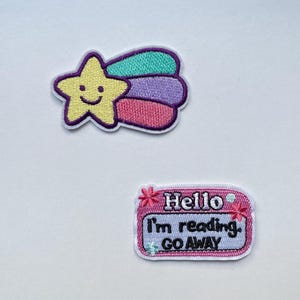 Puede incluir: Dos parches bordados sobre fondo blanco. Uno presenta una estrella amarilla con una cara sonriente y una estela de arcoíris. El otro es un rectángulo rosa con el texto "Hello I'm reading. GO AWAY."