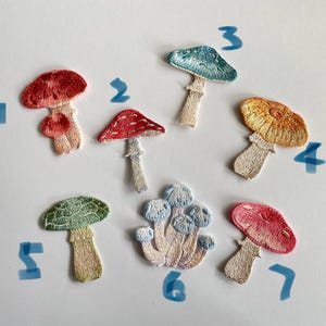 May include: A collection of seven embroidered mushroom patches in various colors. The patches feature detailed stitching and include shades of red, blue, yellow, and green. Each mushroom has a unique design, with varying cap shapes and stem styles.