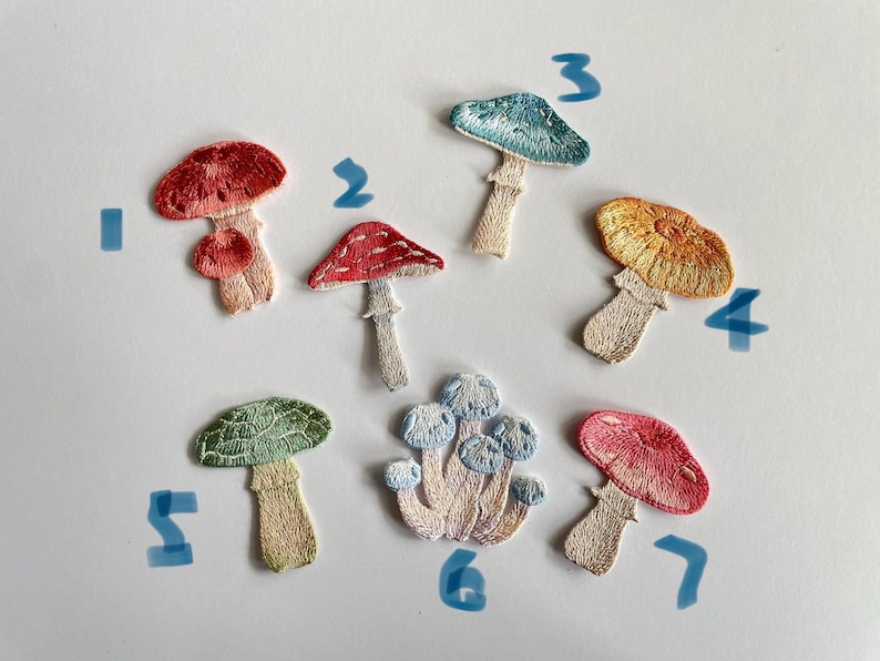 May include: A collection of seven embroidered mushroom patches in various colors. The patches feature detailed stitching and include shades of red, blue, yellow, and green. Each mushroom has a unique design, with varying cap shapes and stem styles.