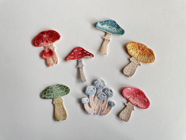 May include: Embroidered mushroom patches in various colors, including red, blue, yellow, and green. The patches feature detailed stitching and are arranged on a white surface. These patches are ideal for adding a whimsical touch to clothing or accessories.