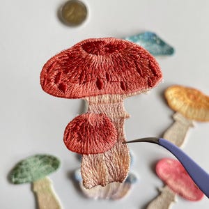 May include: Embroidered mushroom patches in various colors, including red, green, pink, and yellow. The patches are detailed with textured stitching and are in a variety of sizes. A pair of tweezers is holding one of the patches.