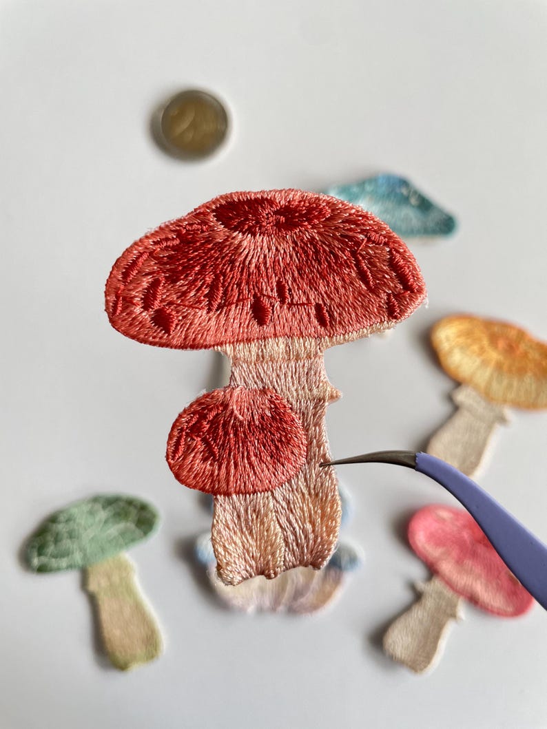 May include: Embroidered mushroom patches in various colors, including red, green, pink, and yellow. The patches are detailed with textured stitching and are in a variety of sizes. A pair of tweezers is holding one of the patches.