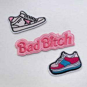 May include: Three embroidered patches: a white sneaker with pink detailing and a star, a pink patch with the words "Bad Bitch", and a sneaker with pink, blue, and white details.