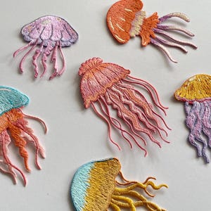May include: Embroidered jellyfish patches in various colors, including pink, orange, yellow, and blue. Each patch features detailed stitching, with flowing tentacles and a rounded bell shape. These patches are suitable for adding to clothing or accessories.