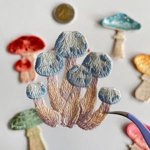 May include: Embroidered mushroom patches in various colors, including red, blue, green, and peach. The central patch features a cluster of mushrooms with blue caps and pink and peach stems. A coin is visible in the background.