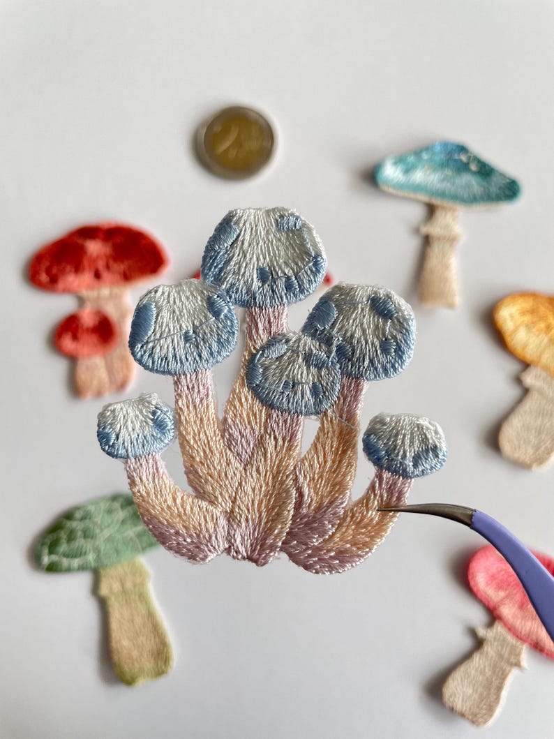 May include: Embroidered mushroom patches in various colors, including red, blue, green, and peach. The central patch features a cluster of mushrooms with blue caps and pink and peach stems. A coin is visible in the background.