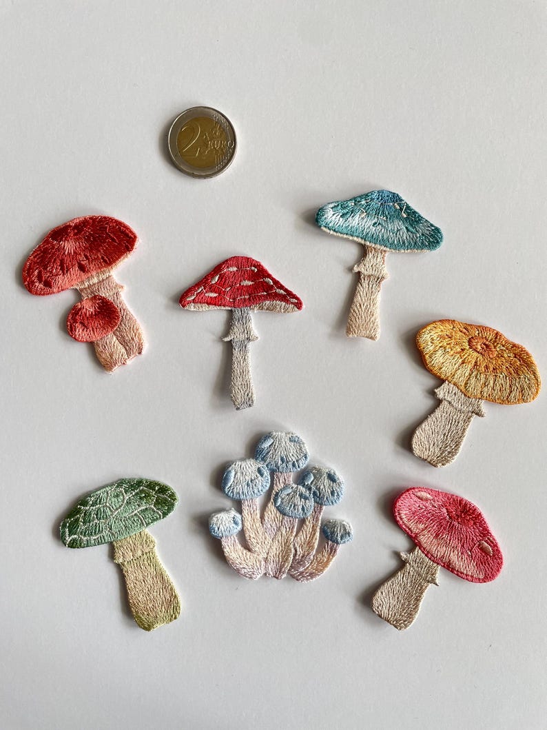 May include: Embroidered mushroom patches in various colors, including red, blue, yellow, and green. The patches feature detailed stitching and are arranged on a white surface. A 2 Euro coin is also present.