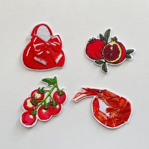 Puede incluir: Cuatro parches bordados: un bolso rojo con un lazo, dos granadas, un racimo de tomates y un camarón. Cada parche tiene un borde blanco sobre un fondo blanco.