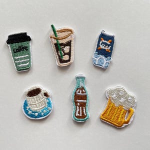 Peut inclure: Écussons brodés représentant des boissons. On y trouve une tasse de café verte, un café glacé, une boisson bleue et blanche, une tasse de café, une bouteille de soda et une chope de bière. Les patchs sont colorés et détaillés.