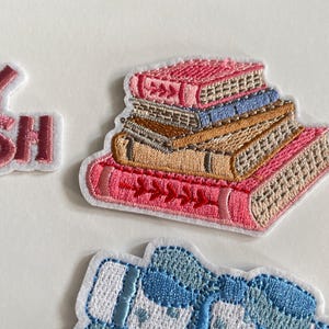 May include: Embroidered patch showing a stack of books in pink, tan, and blue. The books have detailed stitching and a white outline. The patch is designed for clothing or accessories.