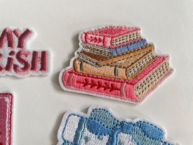 May include: Embroidered patch showing a stack of books in pink, tan, and blue. The books have detailed stitching and a white outline. The patch is designed for clothing or accessories.