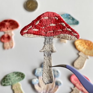 May include: Embroidered mushroom patches in various colors, including red, green, and orange. The central mushroom has a red cap with white spots and a gray stem. A pair of tweezers is holding the mushroom. Other patches are scattered around.