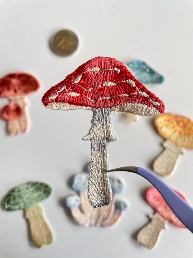 May include: Embroidered mushroom patches in various colors, including red, green, and orange. The central mushroom has a red cap with white spots and a gray stem. A pair of tweezers is holding the mushroom. Other patches are scattered around.