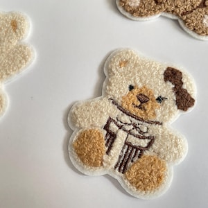 Pode incluir: Close-up de v&aacute;rios patches de ursinhos de pel&uacute;cia. Os patches s&atilde;o branco-sujos com detalhes em castanho nas patas, nariz e uma mancha na cabe&ccedil;a. Um urso tem um cachecol castanho e um presente. Os patches est&atilde;o em uma superf&iacute;cie branca.