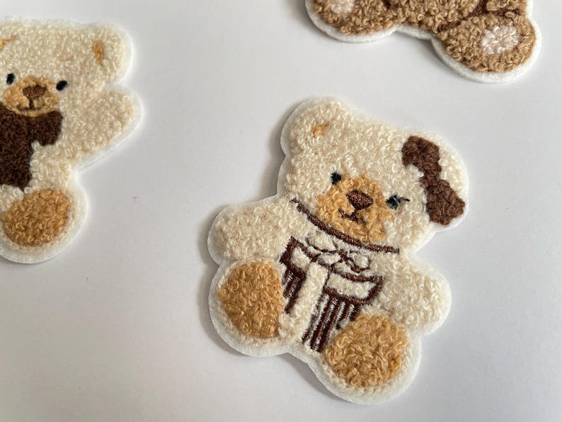 Pode incluir: Close-up de v&aacute;rios patches de ursinhos de pel&uacute;cia. Os patches s&atilde;o branco-sujos com detalhes em castanho nas patas, nariz e uma mancha na cabe&ccedil;a. Um urso tem um cachecol castanho e um presente. Os patches est&atilde;o em uma superf&iacute;cie branca.